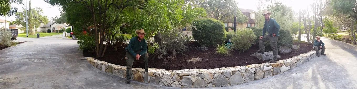 Two people stand next to a rock-lined garden bed in a driveway. Trees and houses are in the background.