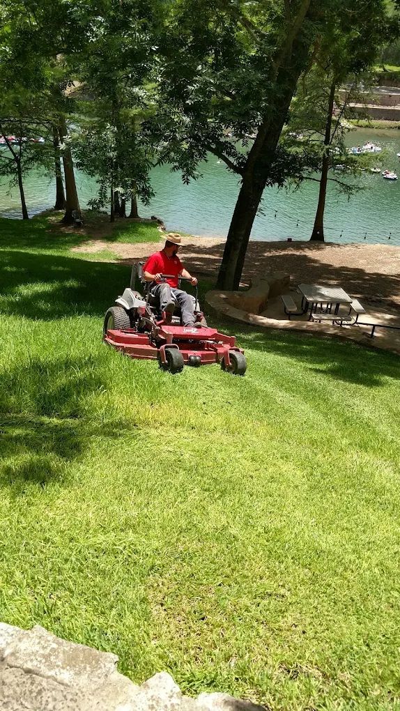 Person mowing a green grassy hill near a lake on a riding mower. Trees and picnic tables are in the background.