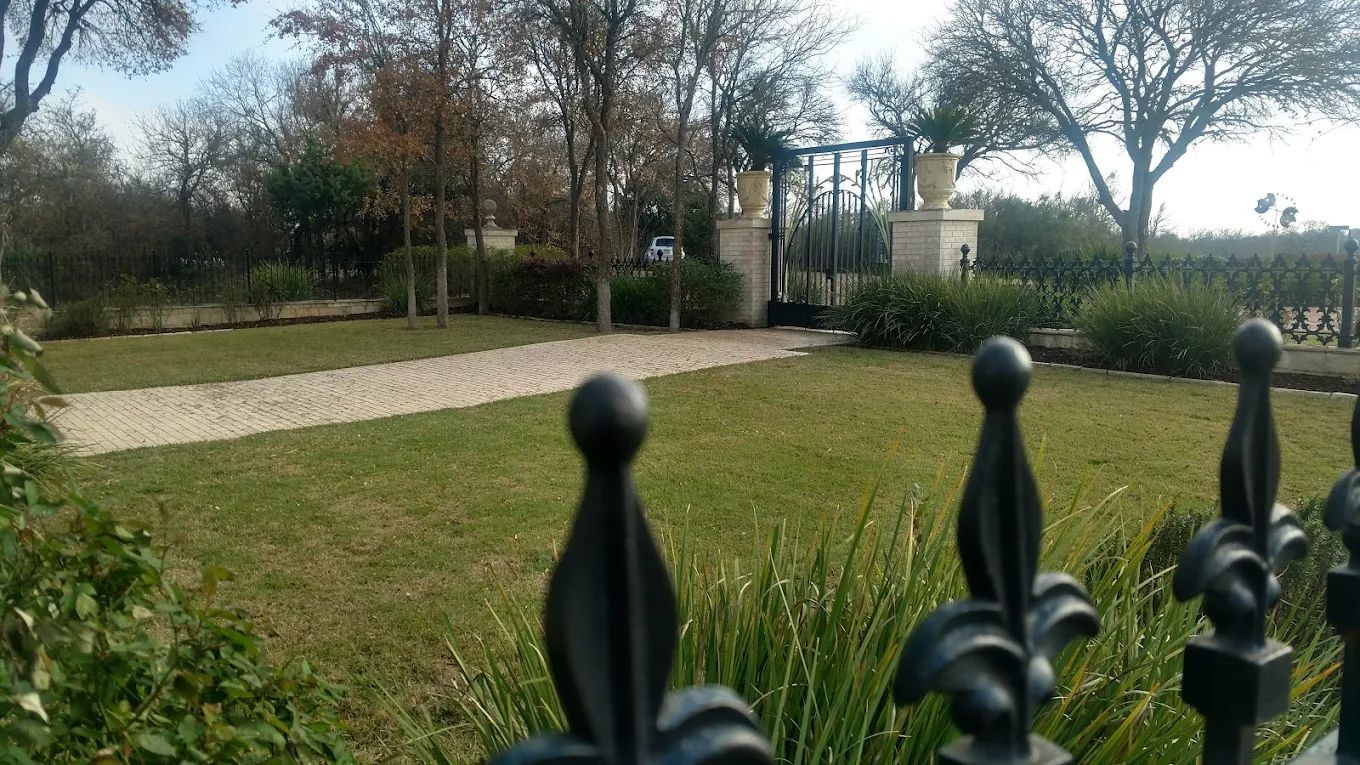 Green lawn with stone path leading to ornate black gate. Black fence in foreground, trees in the background.
