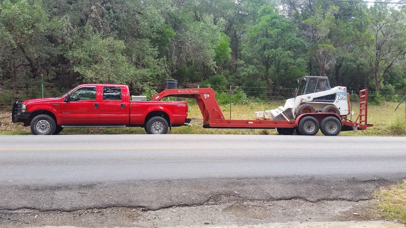 Red truck towing a red trailer with a Bobcat on the side of a paved road, trees in background.