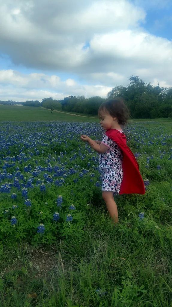 Child in a red cape standing in a field of bluebonnets under a cloudy sky.