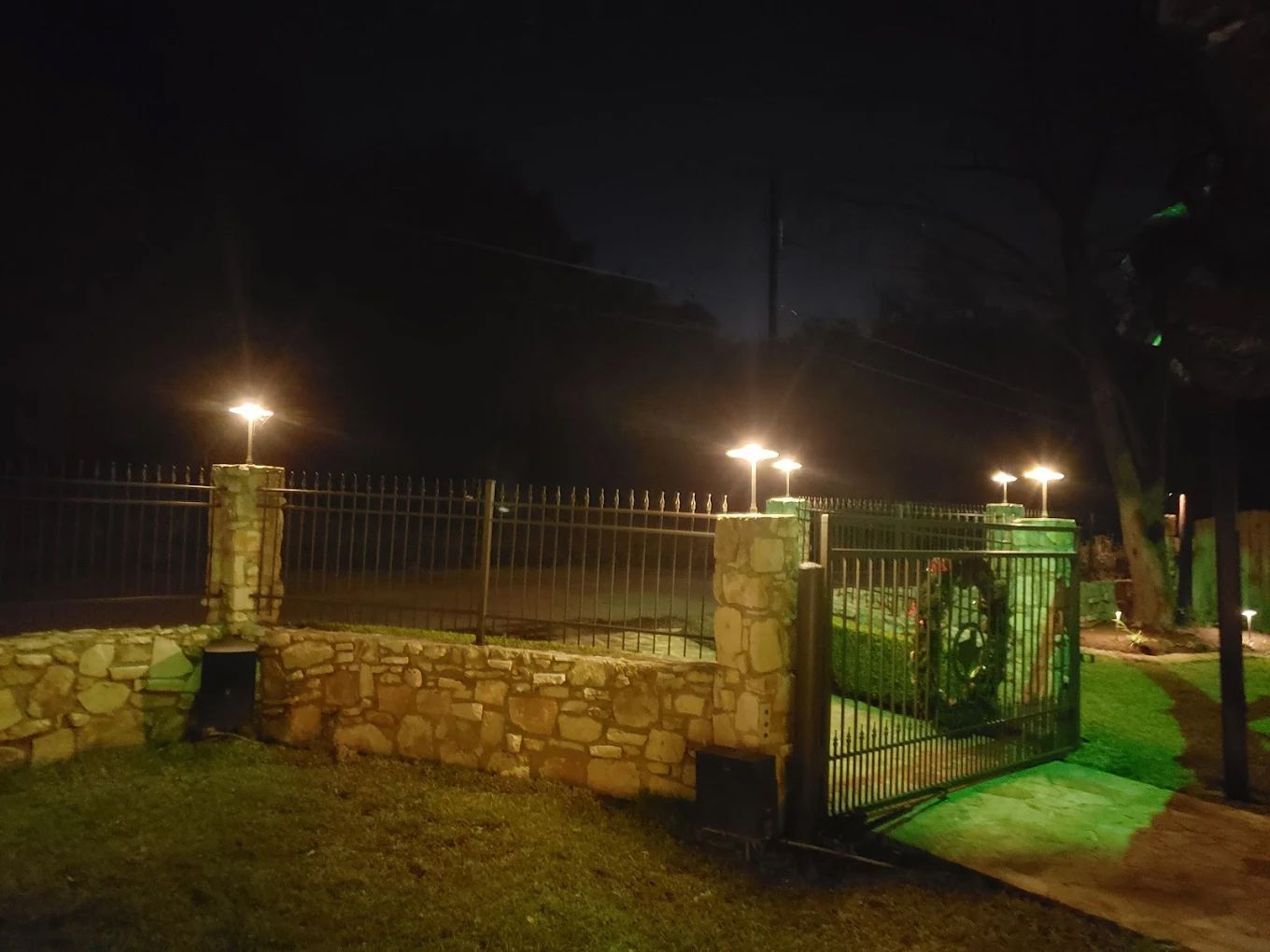 A lit gate and fence at night, with stone pillars and a green lawn.