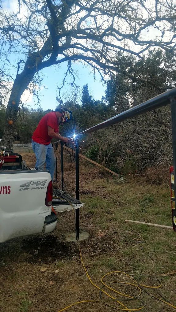 A person welds metal fence posts, standing on a truck bed, outdoors near trees.