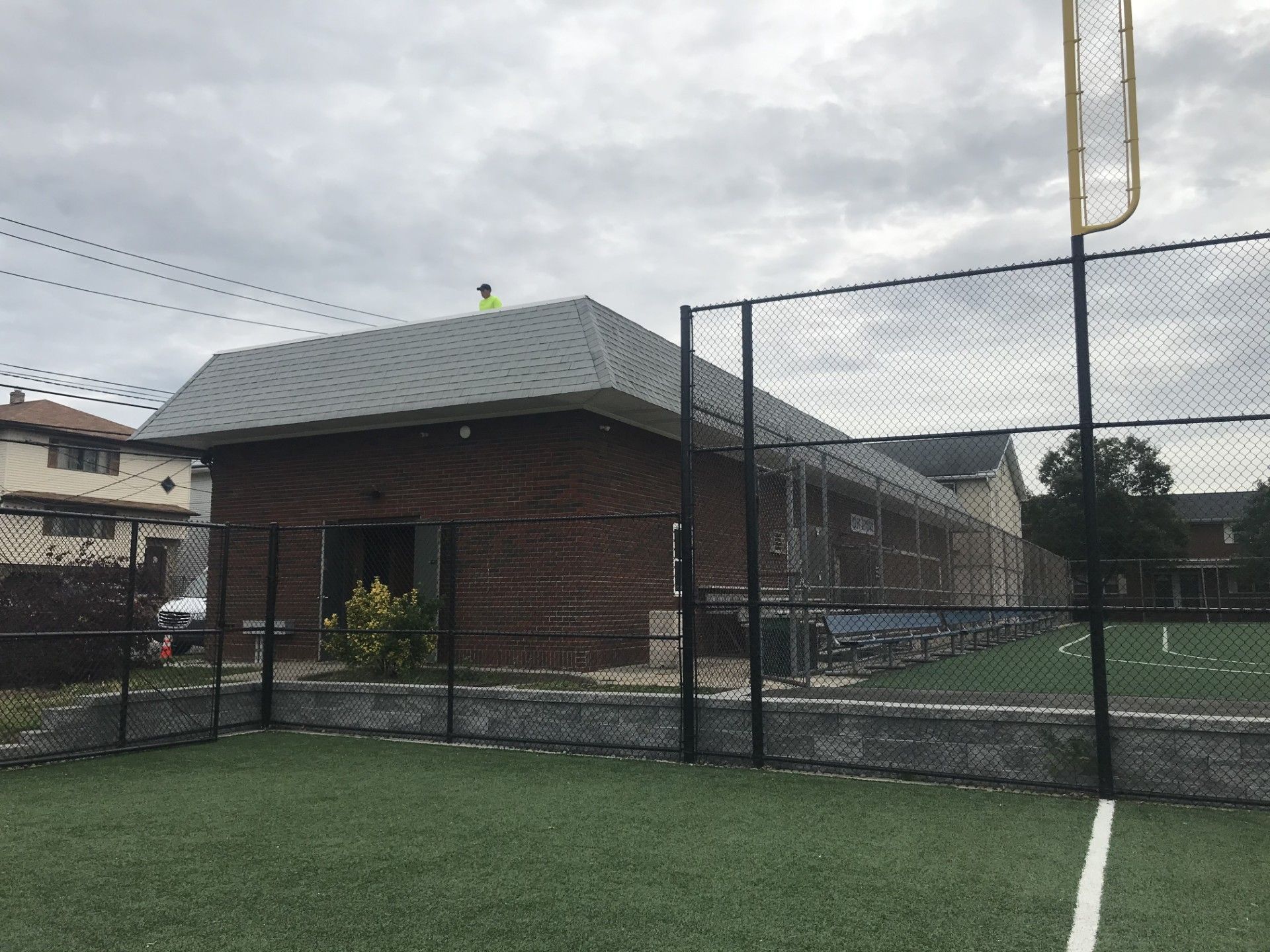 A person in a neon vest working on the roof of a brick building next to a fenced-in turf field.