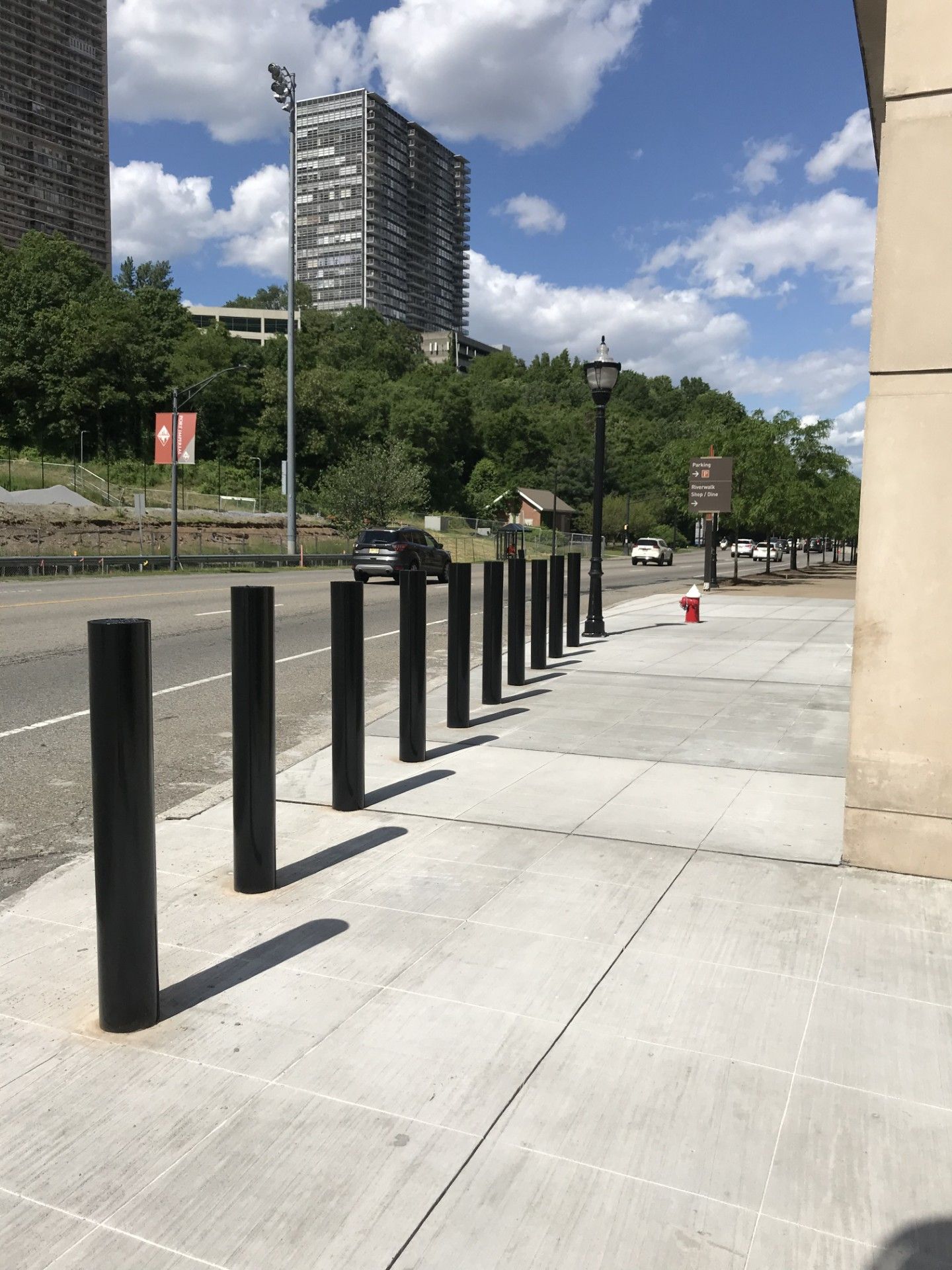 A row of black bollards on a light concrete sidewalk next to a road, with trees and a tall building in the background.