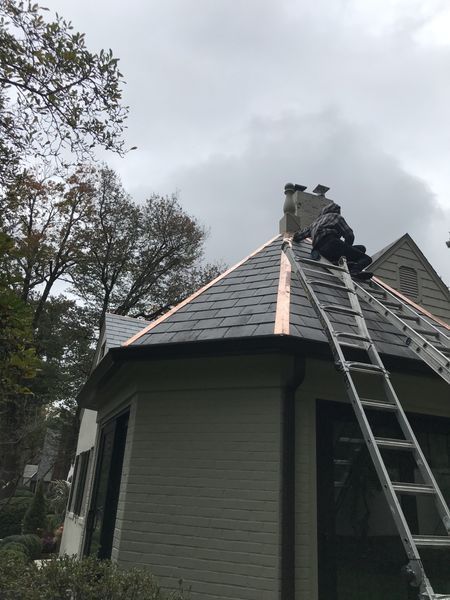 Workers on a roof with a ladder, installing a dark slate roof with copper flashing, cloudy sky.