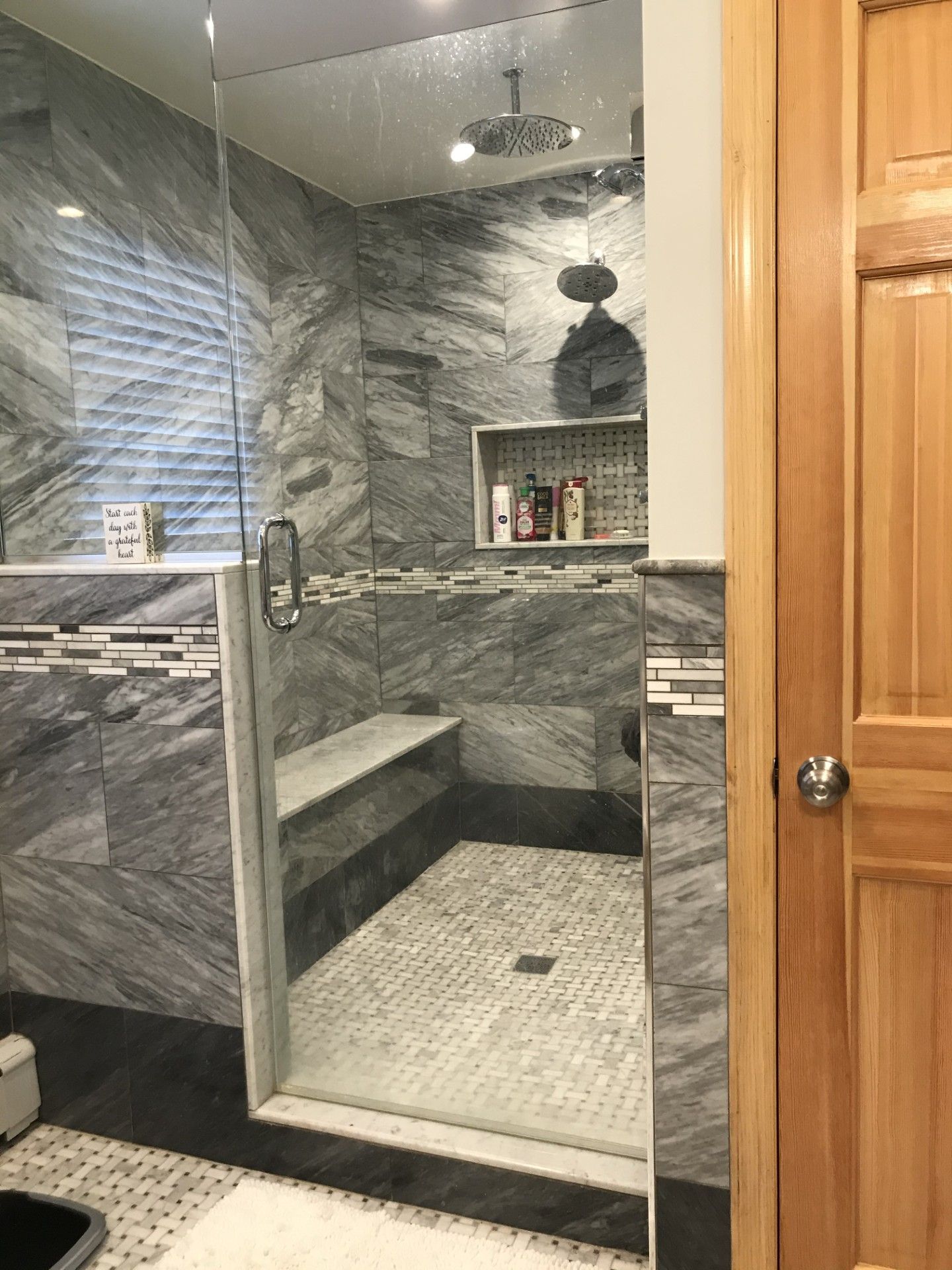 Modern walk-in shower with gray marbled tile, a glass door, built-in bench, rainfall showerhead, and recessed shelf.