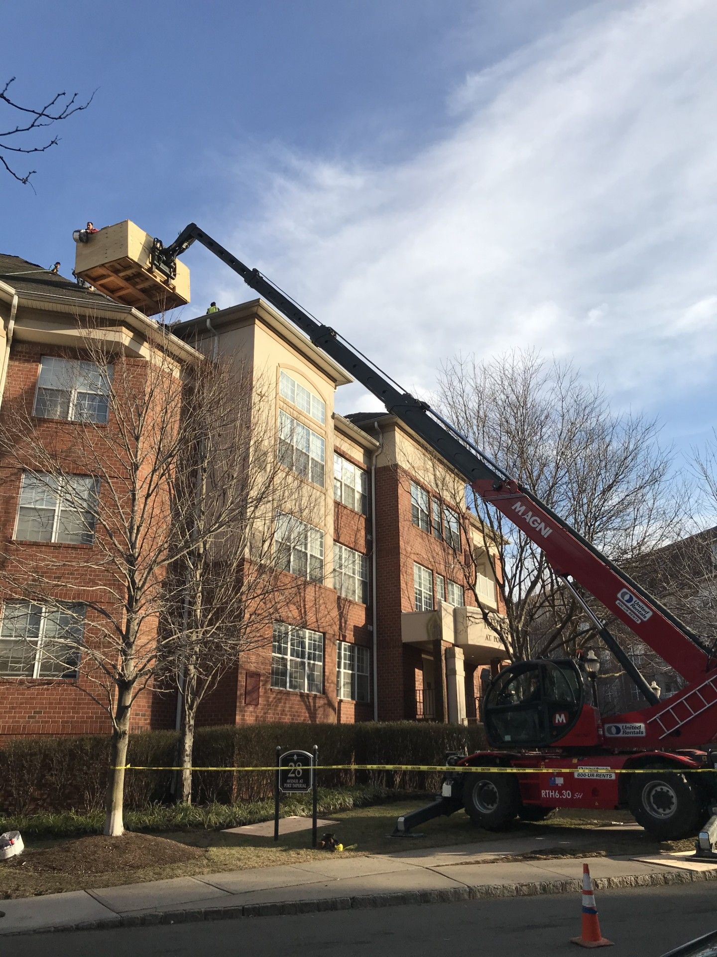 A red construction crane lifts a large wooden crate to the roof of a three-story brick apartment building.