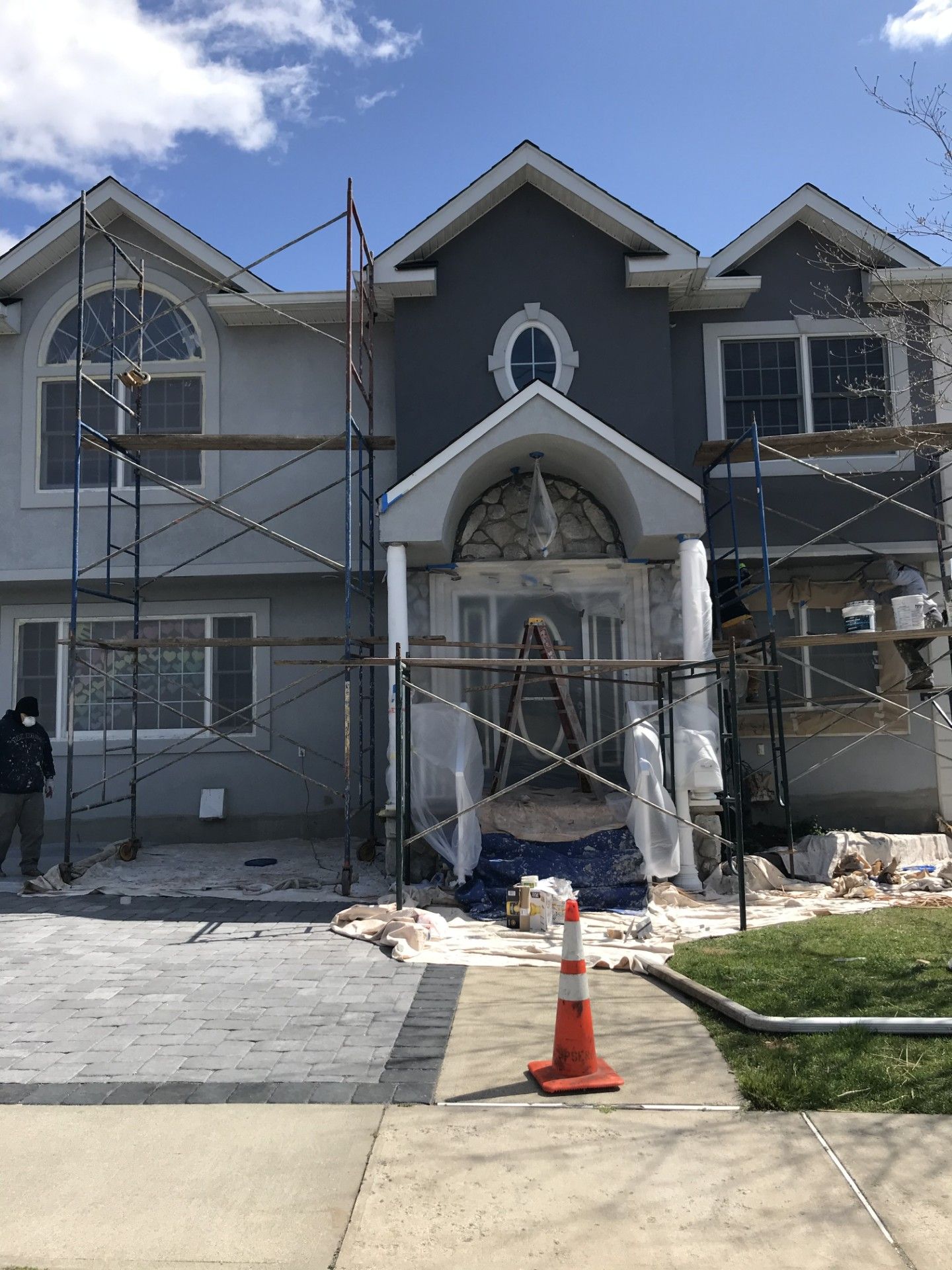 A two-story house under renovation, featuring gray stucco, exterior scaffolding, and a safety cone in the front yard.