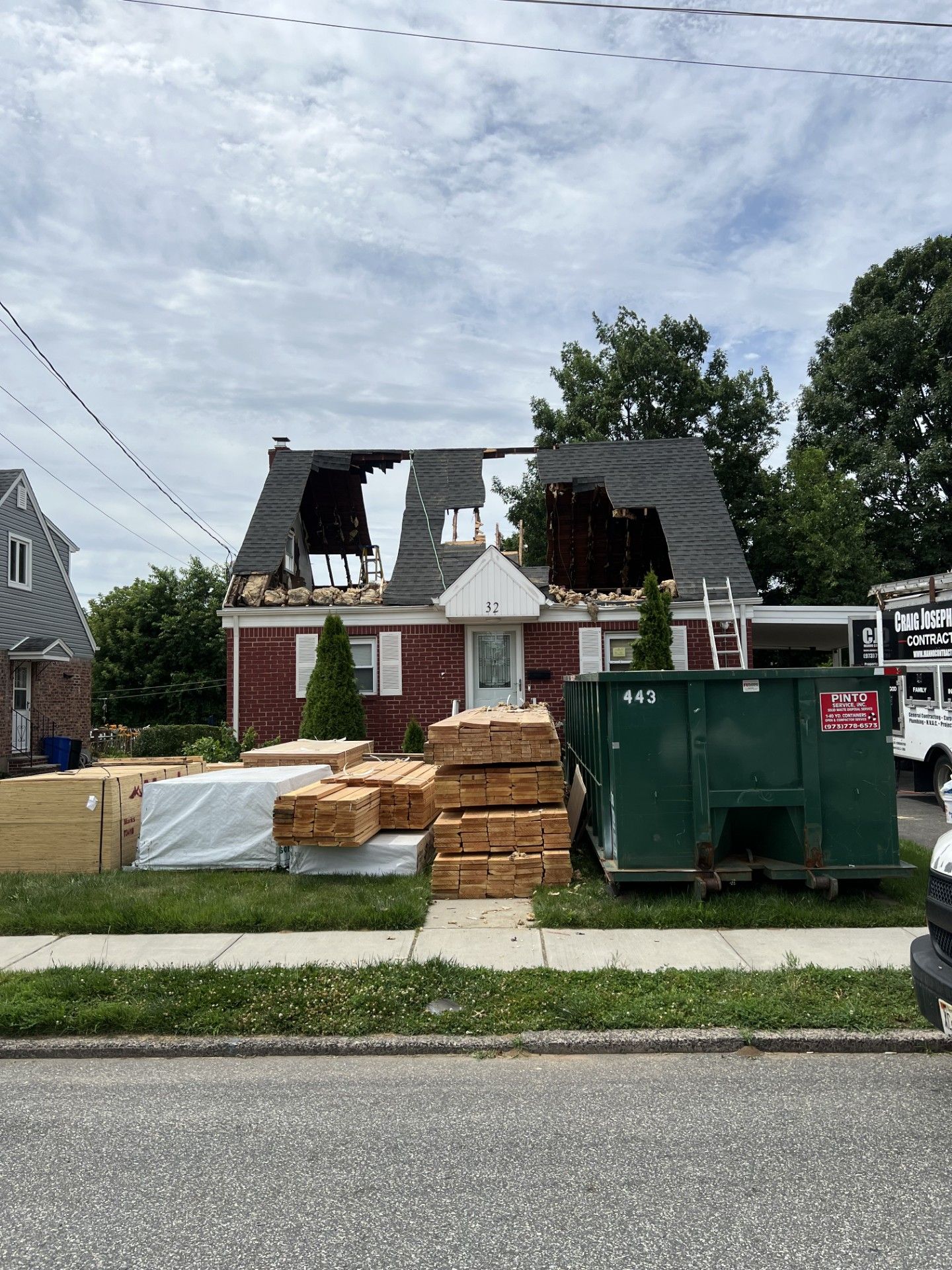 A red brick house with a partially demolished roof, stacks of lumber in the yard, and a large green dumpster.