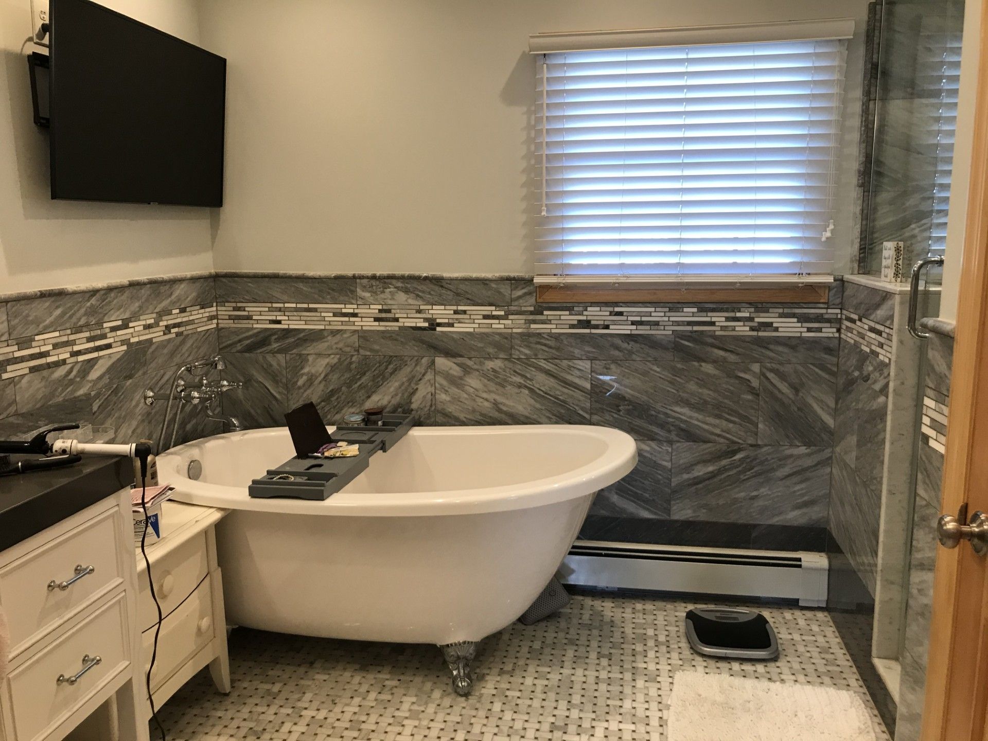 A bathroom featuring a freestanding white tub, grey stone tile walls, a wall-mounted TV, and a white vanity.