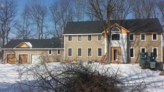 House under construction in a snowy landscape. Tan siding, dark roof, construction equipment.