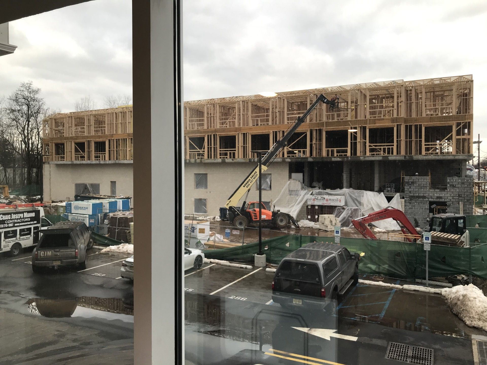 Construction site with a partially built wooden-framed building, heavy machinery, and parked cars under a cloudy sky.