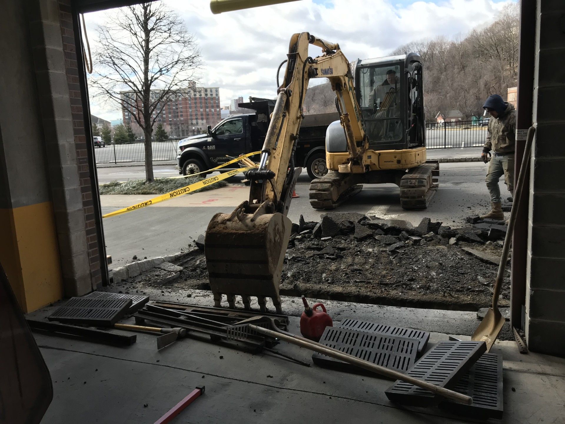 An excavator sits outside an open bay door, with construction tools and grates on the floor in the foreground.