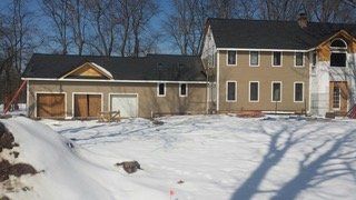 House under construction in a snowy landscape. Tan stucco exterior, black roof, and garage doors.