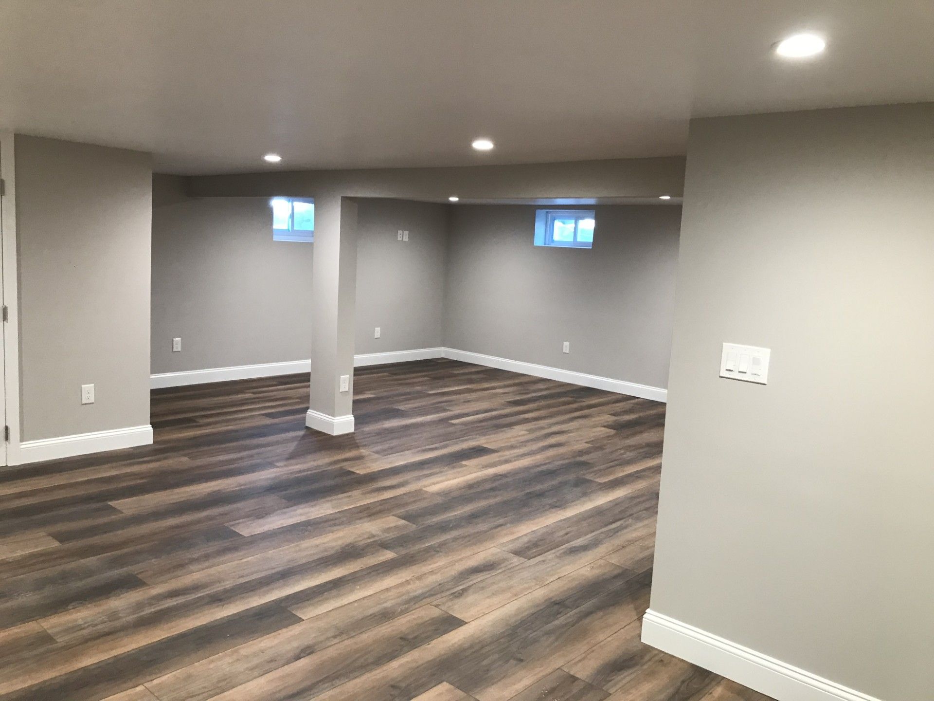 A renovated basement with gray walls, white trim, recessed lighting, and wood-look flooring featuring a support pillar.