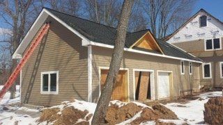 Tan building under construction with a black roof, white trim, and a red ladder. Snow on ground.