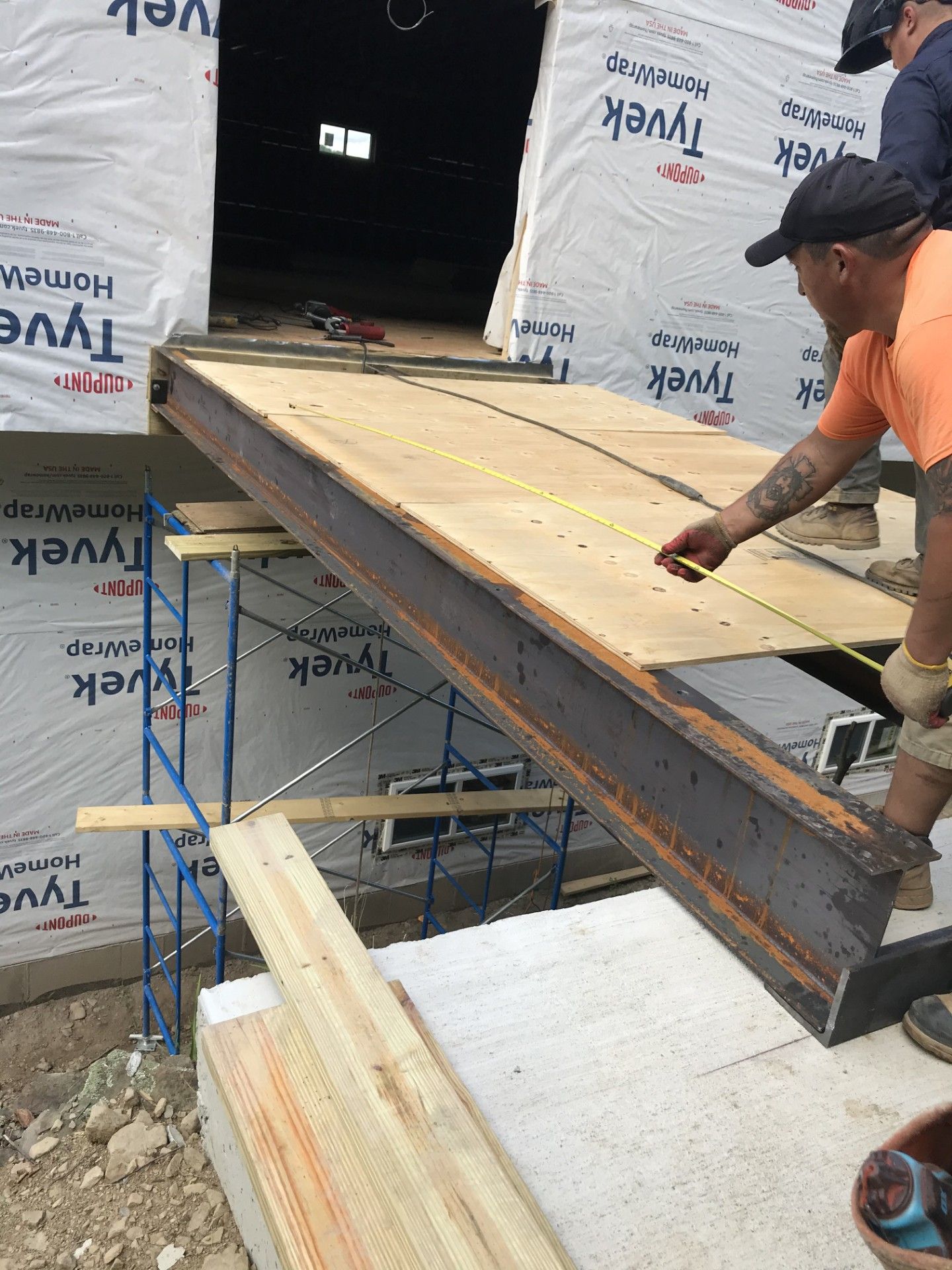 A construction worker uses a yellow tape measure to gauge the length of a steel I-beam supported by scaffolding.