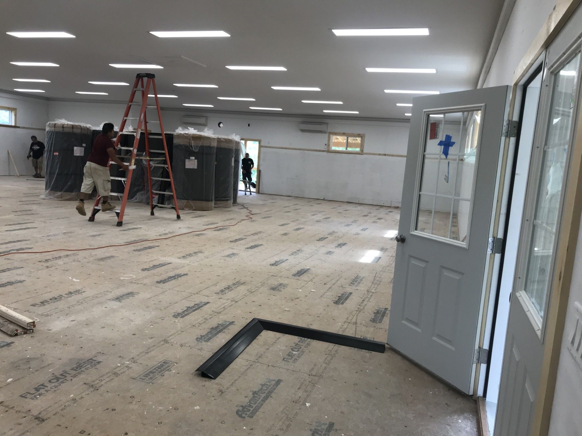 Two workers in a large, indoor construction site with a subfloor, lighting fixtures, and industrial equipment.
