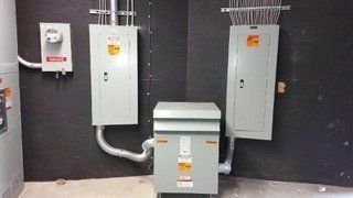 Electrical equipment in an industrial setting. Meter, two electrical panels, and transformer mounted on a black wall.