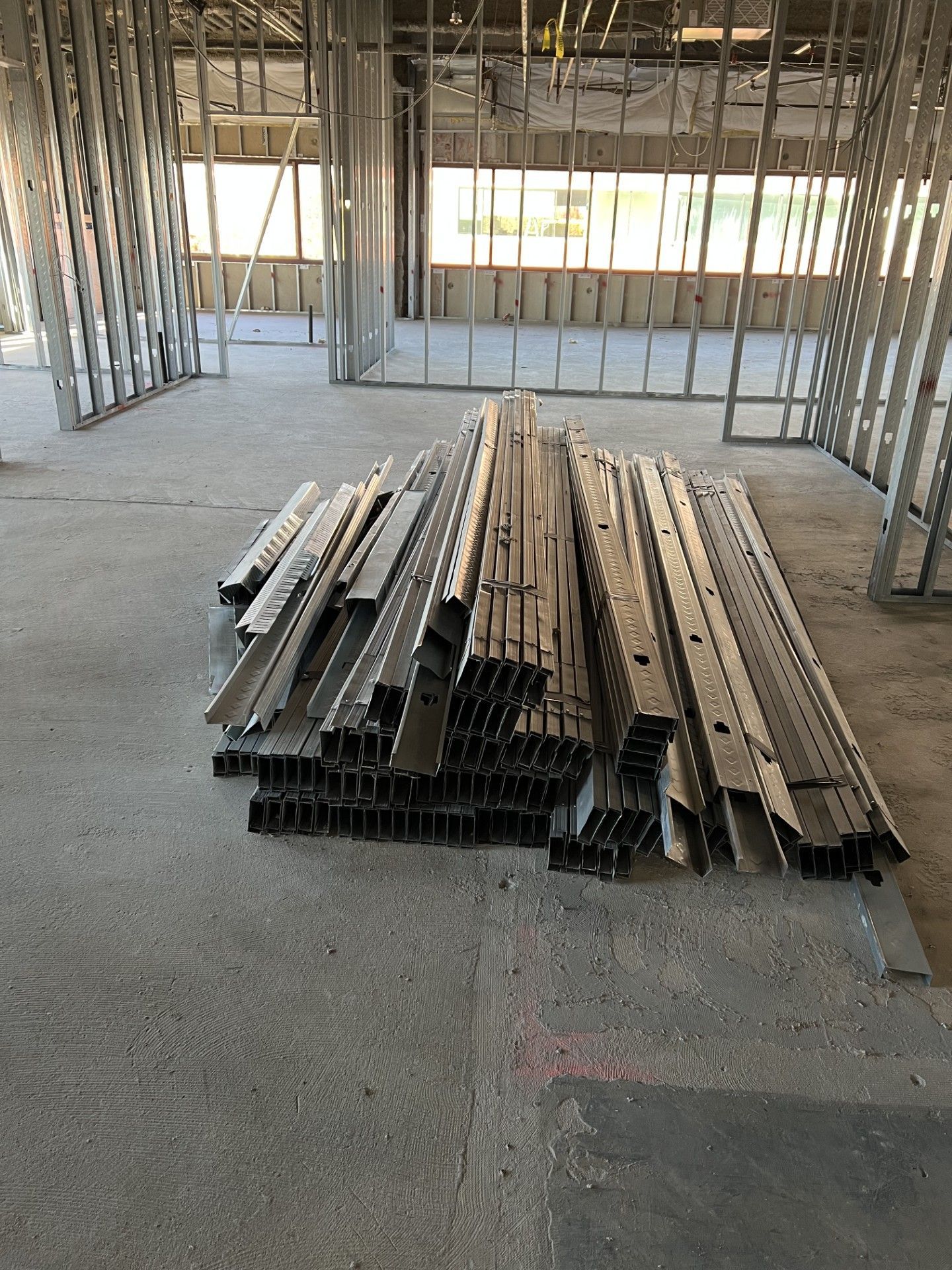 A stack of metal steel studs lies on the concrete floor of a building under construction, with wall framing in the back.