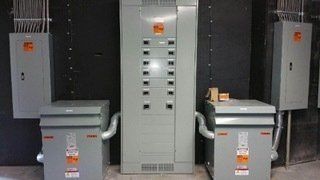 Electrical panel with two transformers and additional boxes on a gray wall.