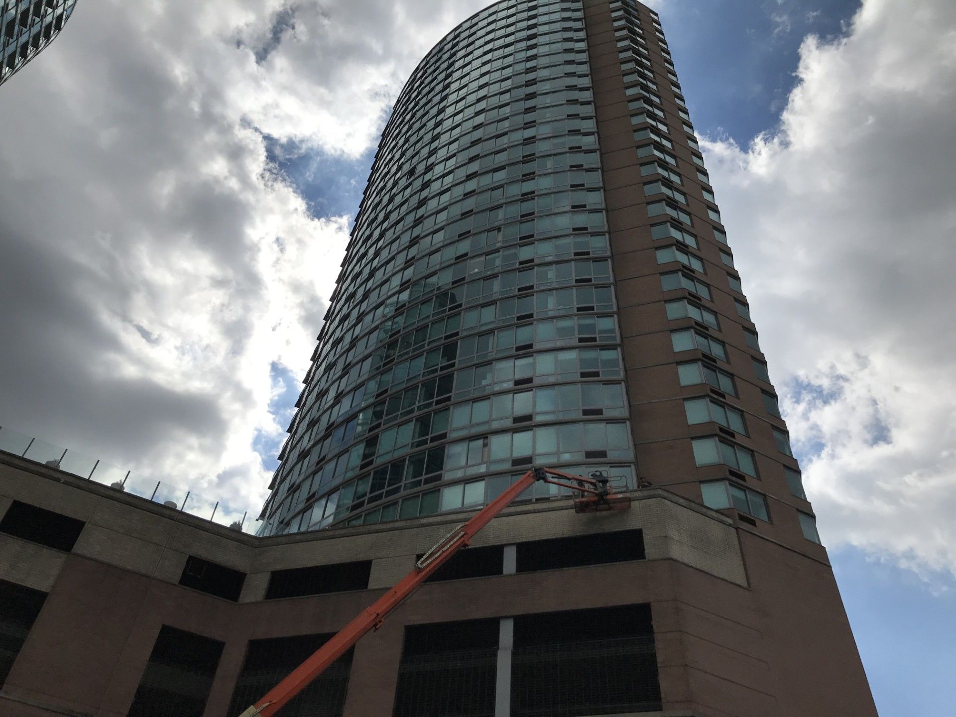 A tall, cylindrical skyscraper with a red-brick base under a cloudy sky, seen from a low angle with a boom lift extending.