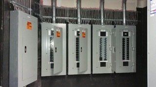 Four open electrical panels, gray metal, installed on a wall with metal conduits above. A closed panel is on the left.