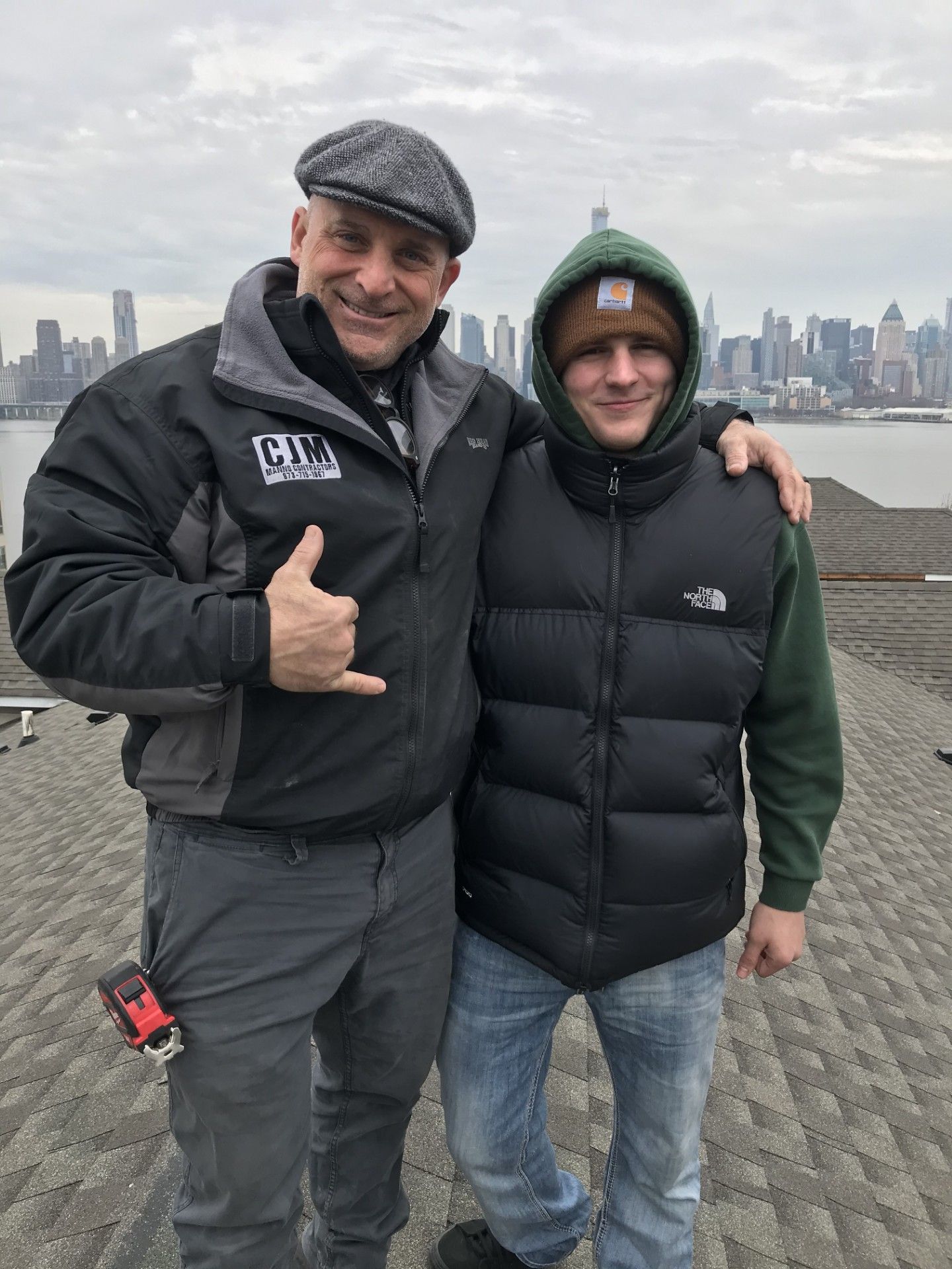 Two people posing for a photo outdoors, one wearing a gray cap and jacket, the other a beanie and vest, with a city skyline.