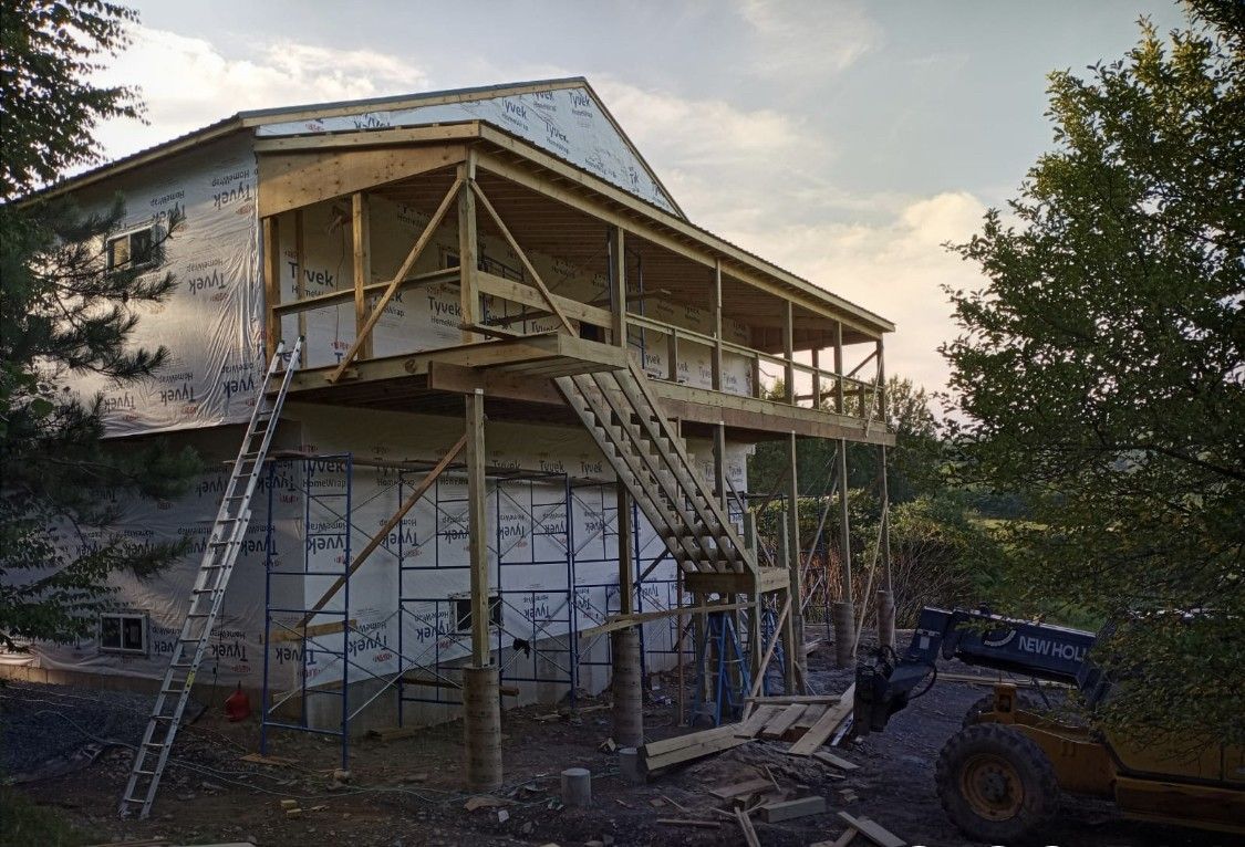 A two-story house under construction with a large wooden deck and outdoor staircase being built, viewed from the yard.