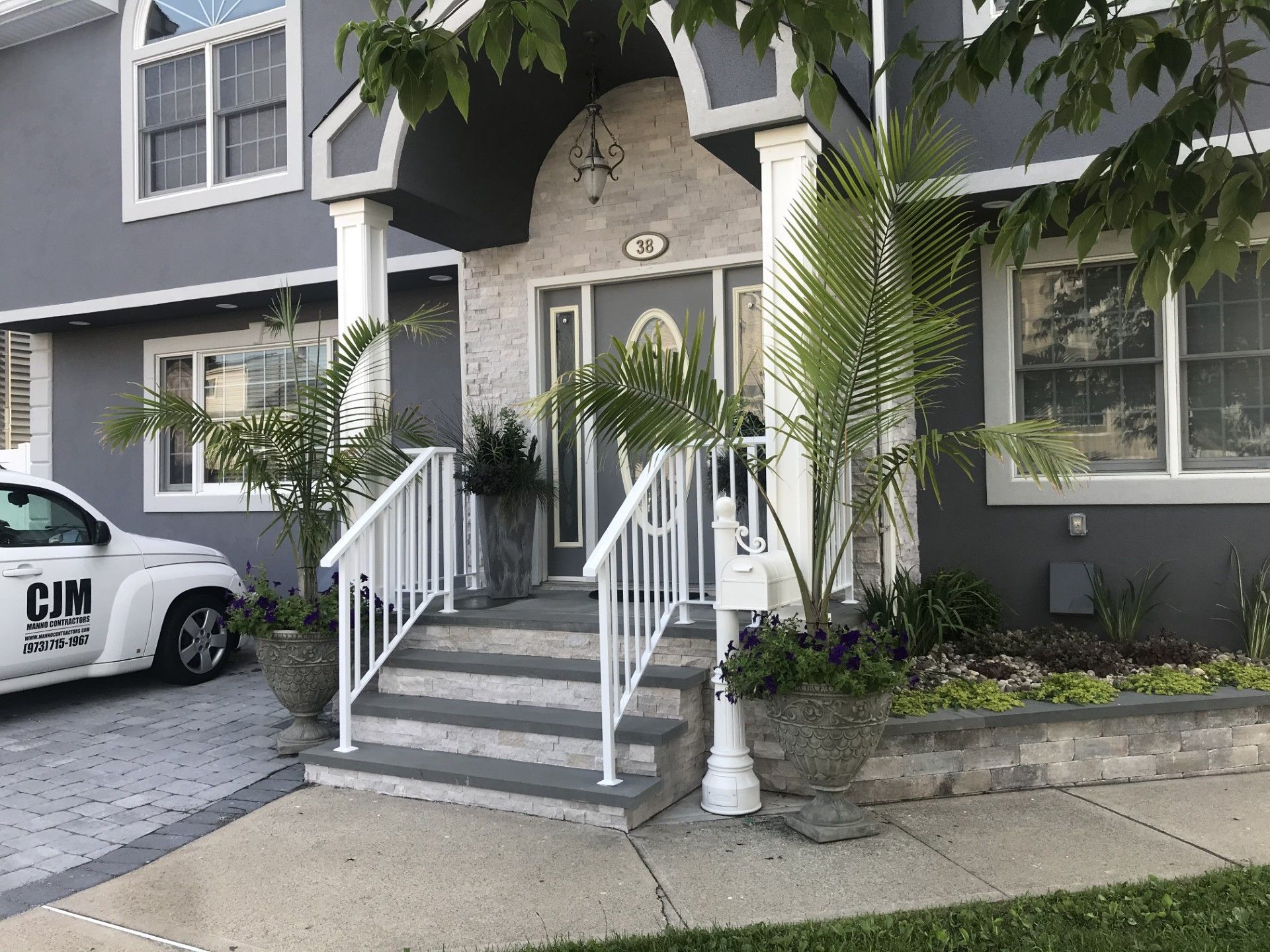 A gray house with a stone-accented front entrance, white railings on stone steps, two potted palms, and a parked car.