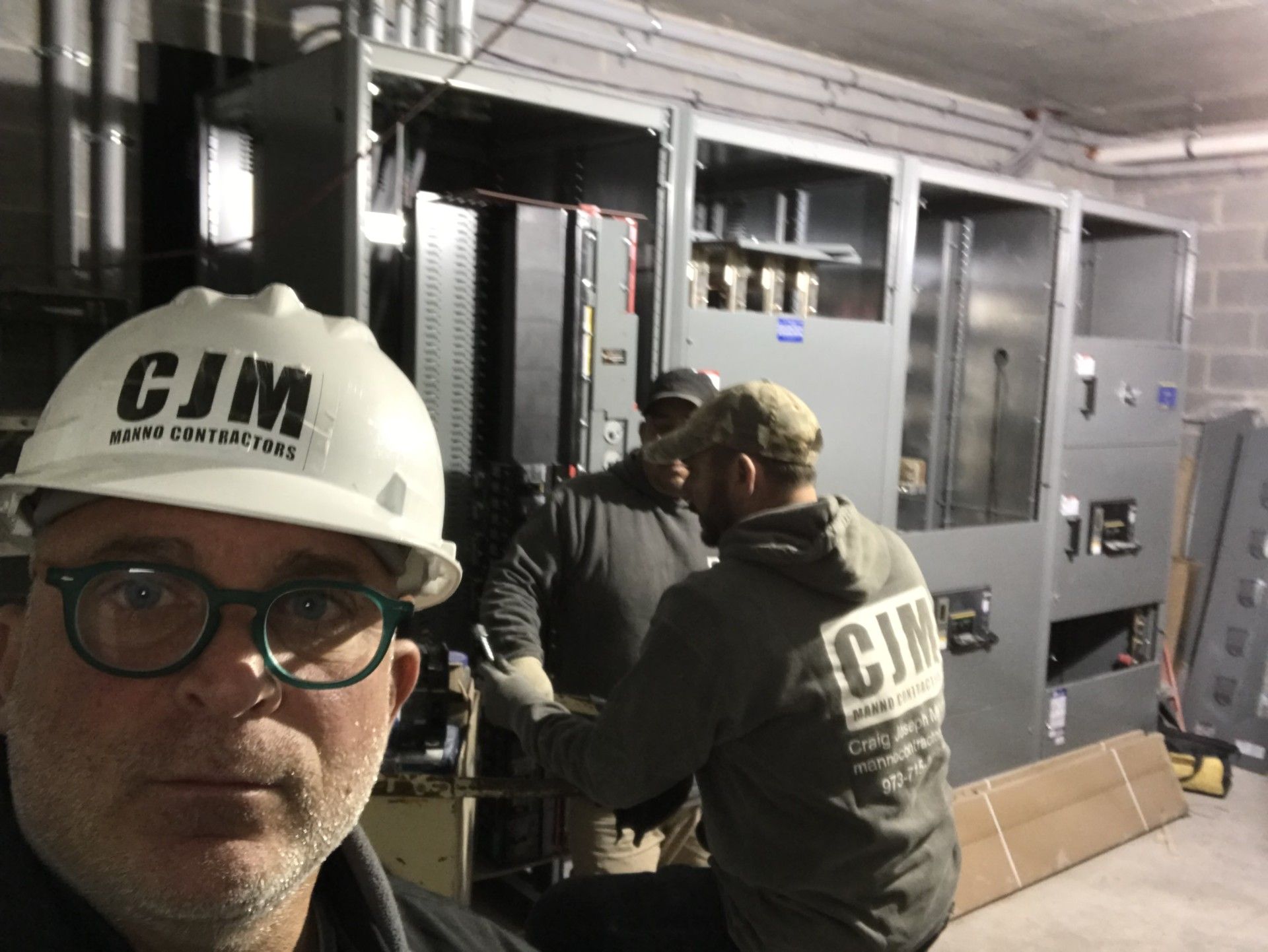 Three workers wearing CJM branding on hard hats and hoodies work on a large electrical control cabinet in a utility room.
