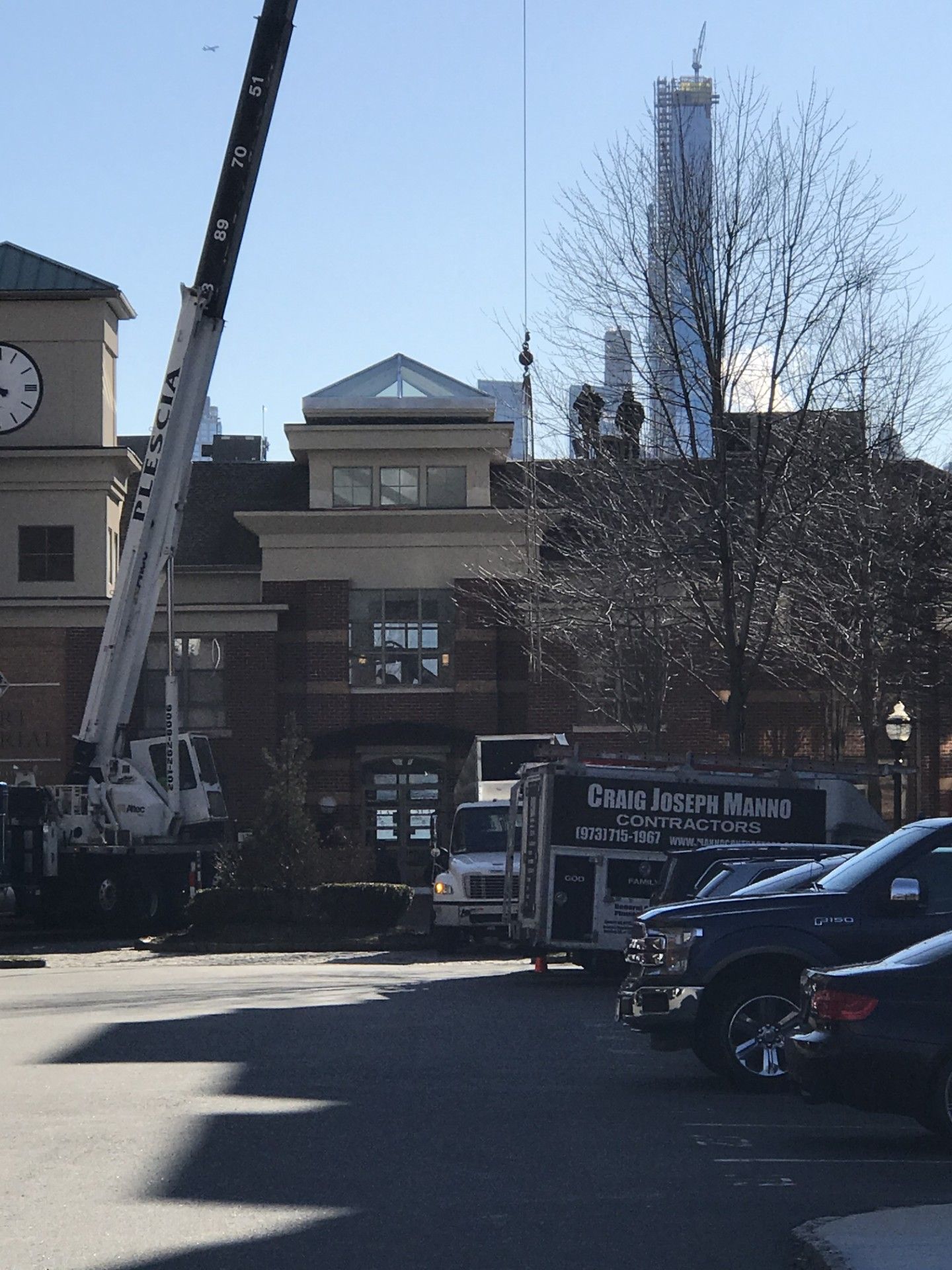 A large crane stands in front of a brick building with a clock tower and a modern high-rise in the background.