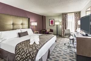 Hotel room with a large bed, TV, desk, armchair, and patterned carpet. Purple and gold color scheme.
