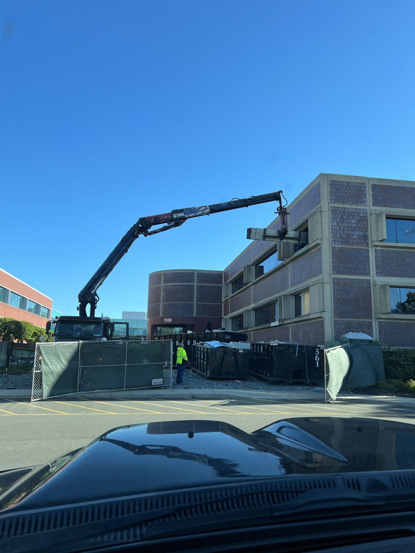 A crane lifts a large construction piece into a window of a multi-story brick building, viewed from a car dashboard.