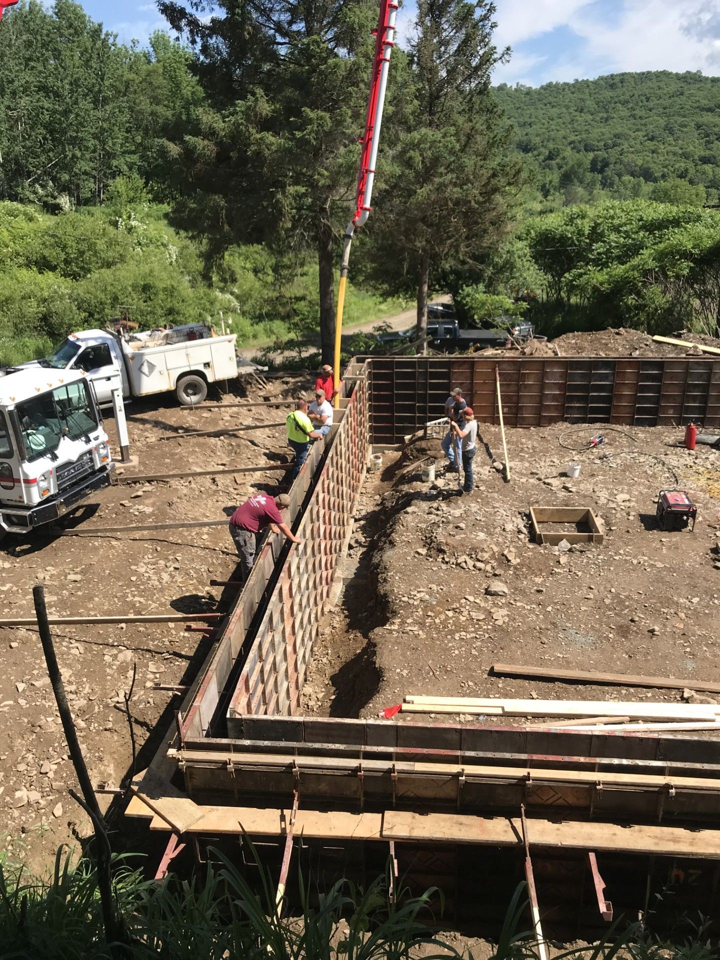 Construction workers assemble metal concrete forms for a building foundation at a sunny, wooded job site.