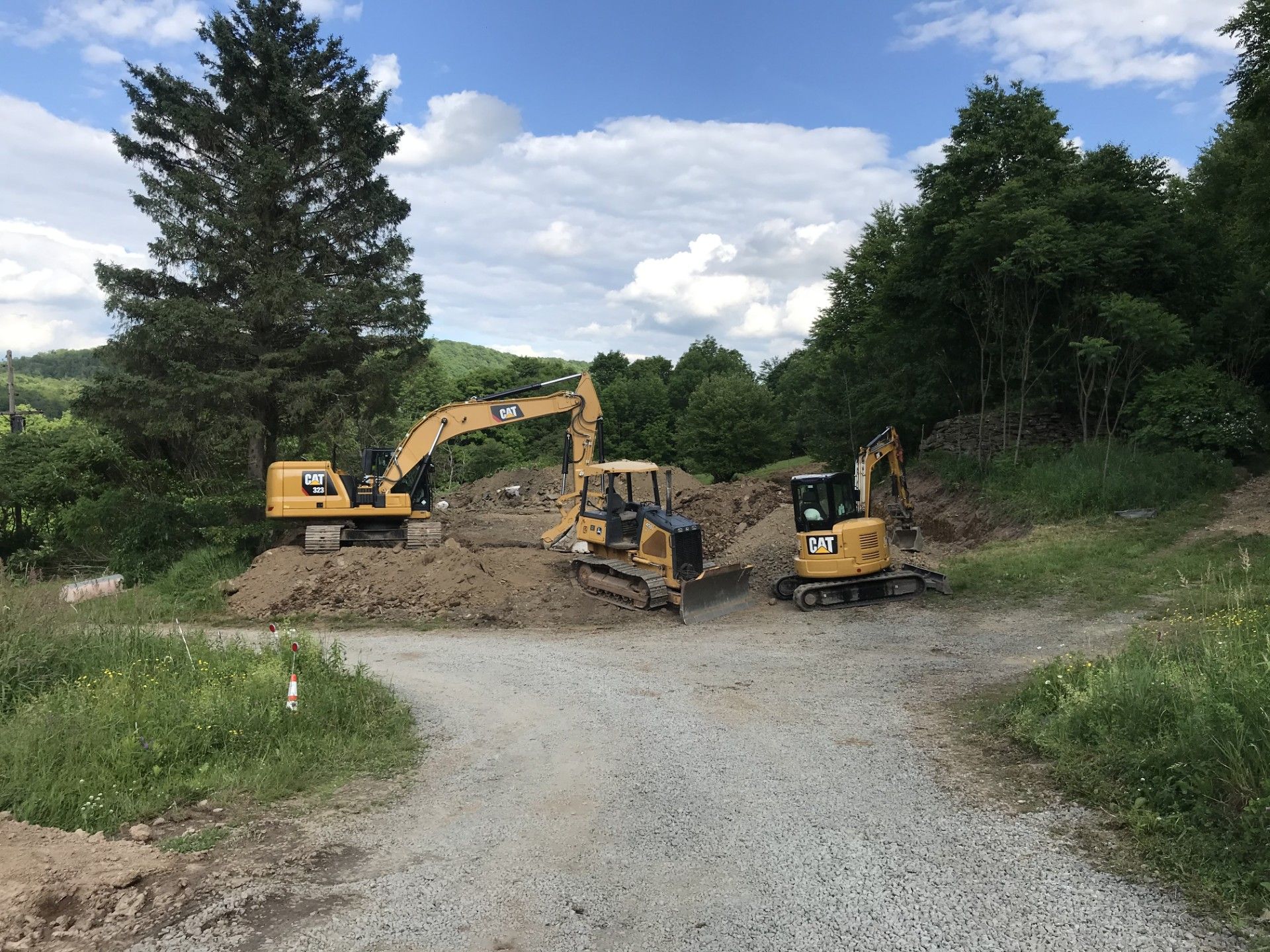 Three yellow construction vehicles are parked on a dirt lot surrounded by trees under a partly cloudy sky.