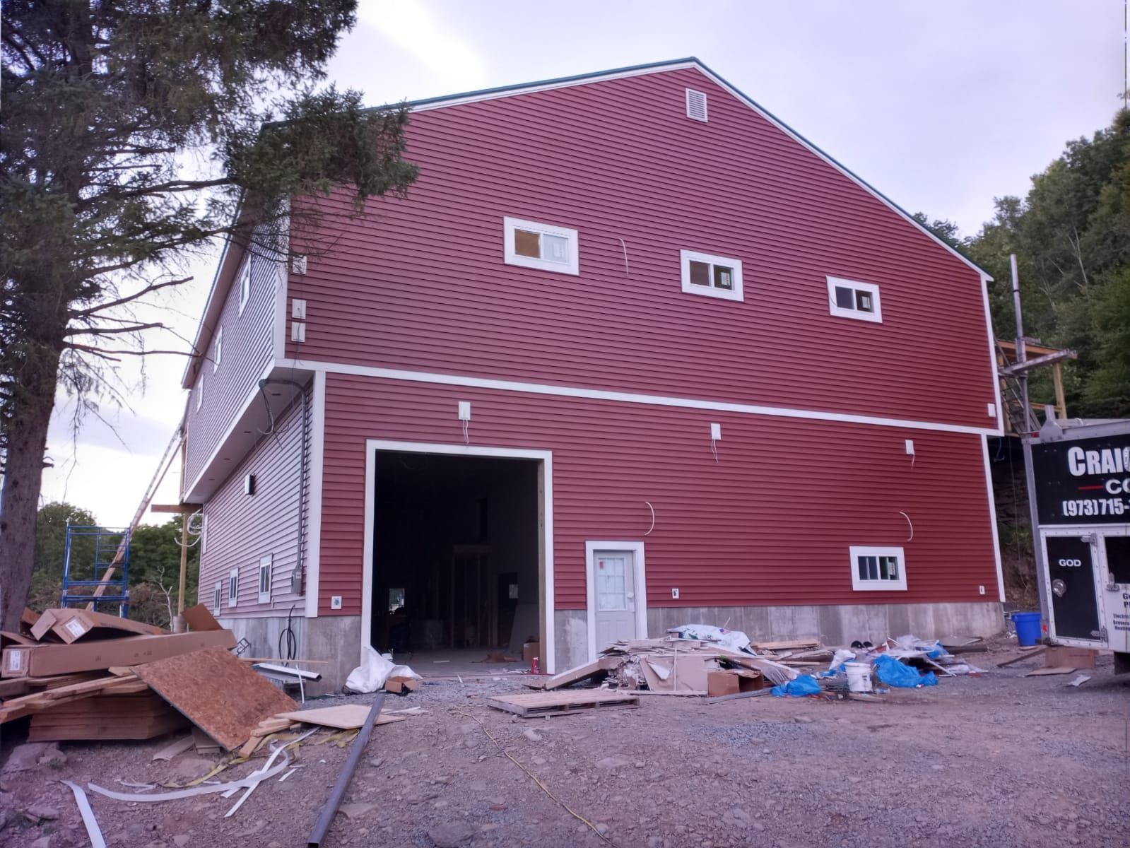 A large, unfinished two-story barn with red horizontal siding and white trim, featuring a garage door and scattered debris.
