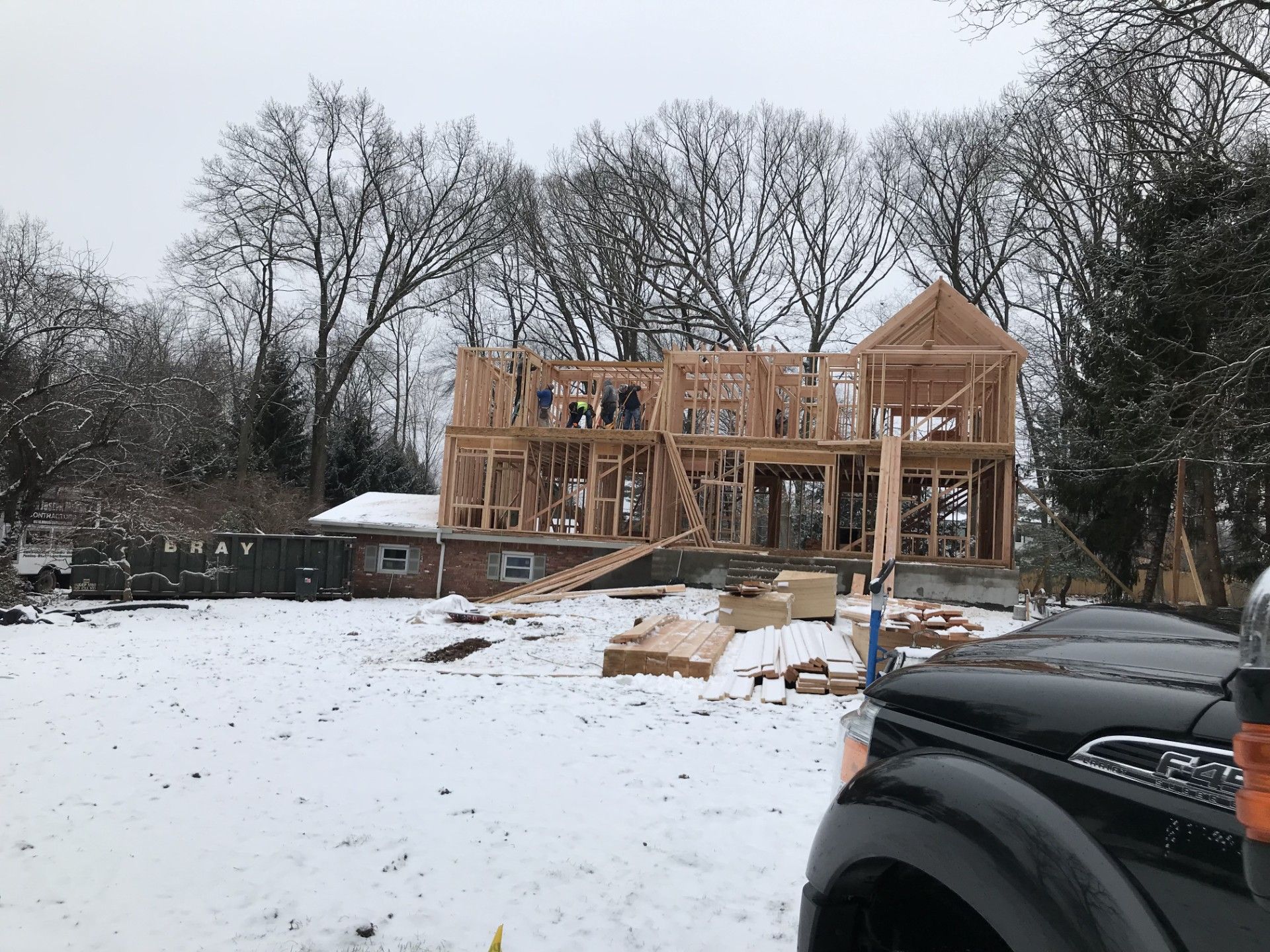 A house under construction with exposed wooden framing stands on a snow-covered lot near a black truck.