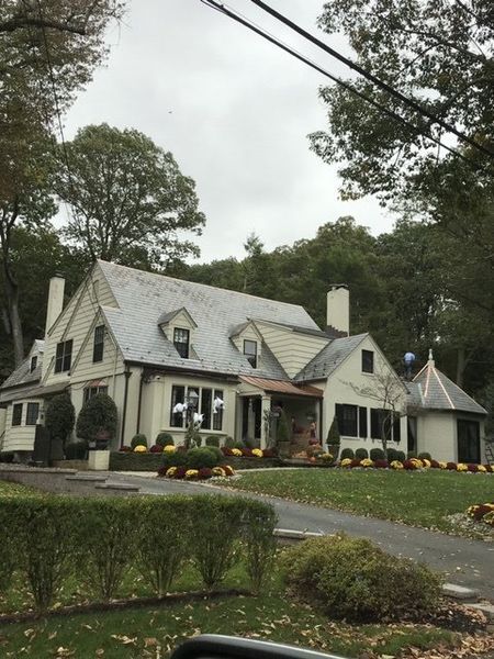 Cream-colored house with dark roof and landscaping, on a tree-lined drive under a cloudy sky.