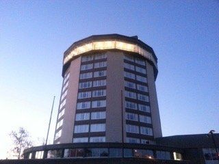 A tall, cylindrical building with a brightly lit top section and many windows against a blue sky.