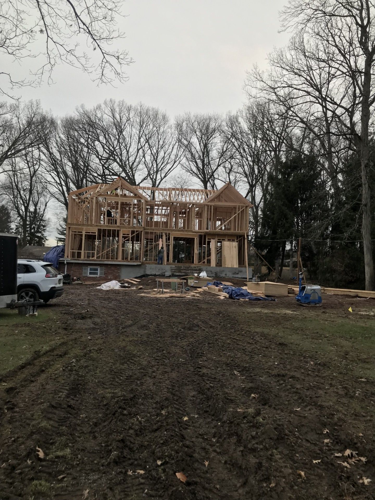 A wooden frame of a two-story house under construction in a muddy, wooded lot on an overcast day.
