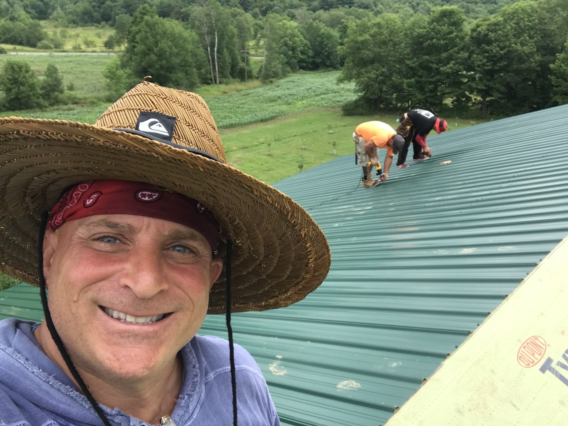 A smiling person in a straw hat and bandana takes a selfie on a green metal roof, with two others working in the distance.