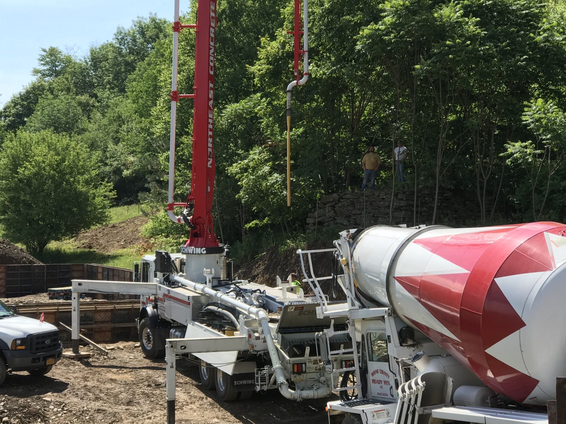 A red and white concrete pump truck and mixing truck stationed at an outdoor construction site with trees in the background.