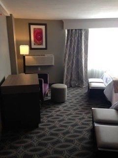 Hotel room with a bed, desk, chair, ottoman, patterned carpet, and curtains.