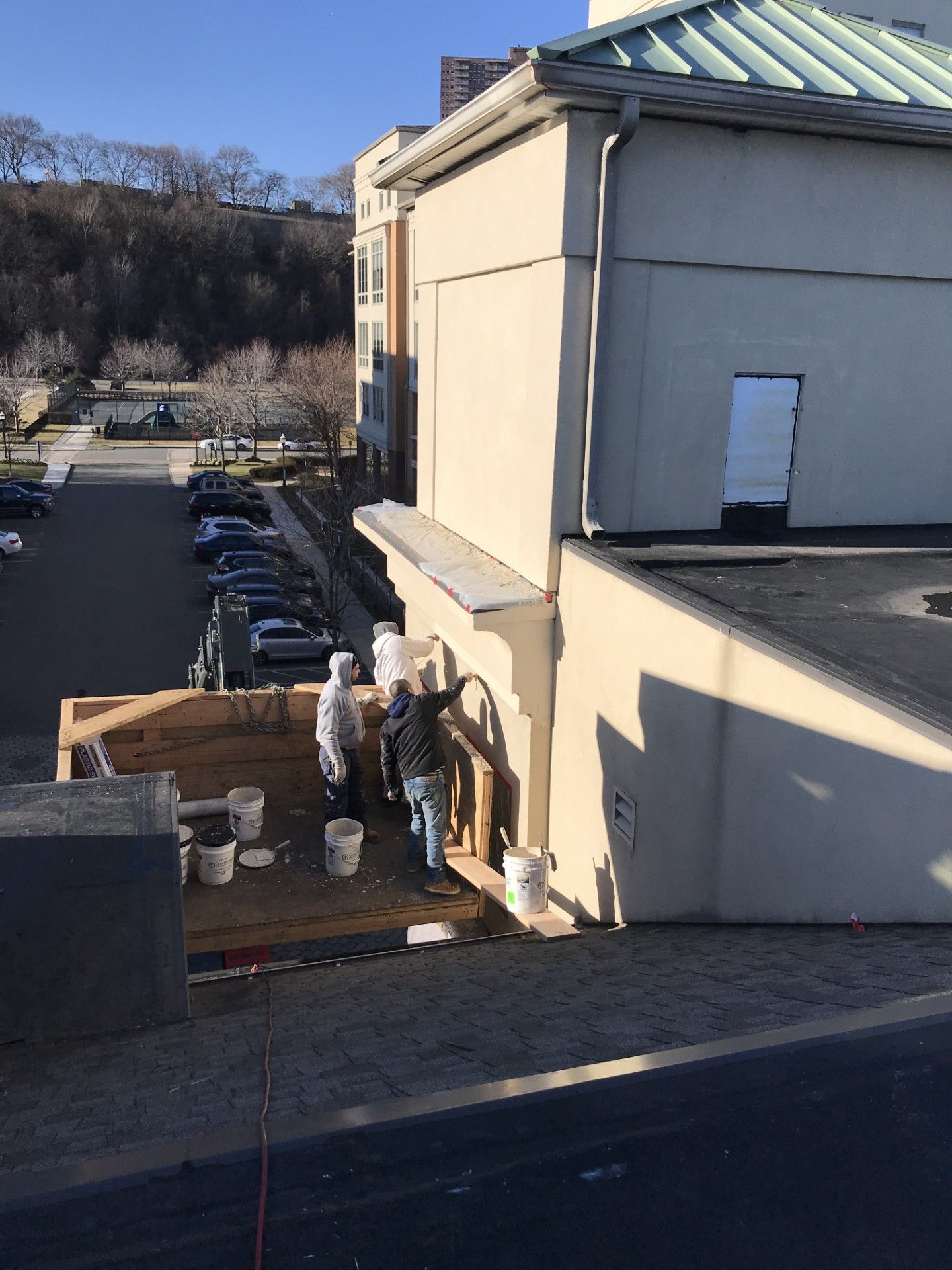 Two people in work clothes repair a beige building wall while standing on a wooden scaffold above a parking lot.