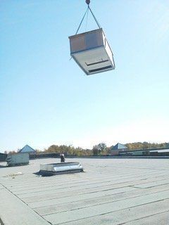A rooftop HVAC unit being lifted by a crane, above a flat commercial roof on a sunny day.
