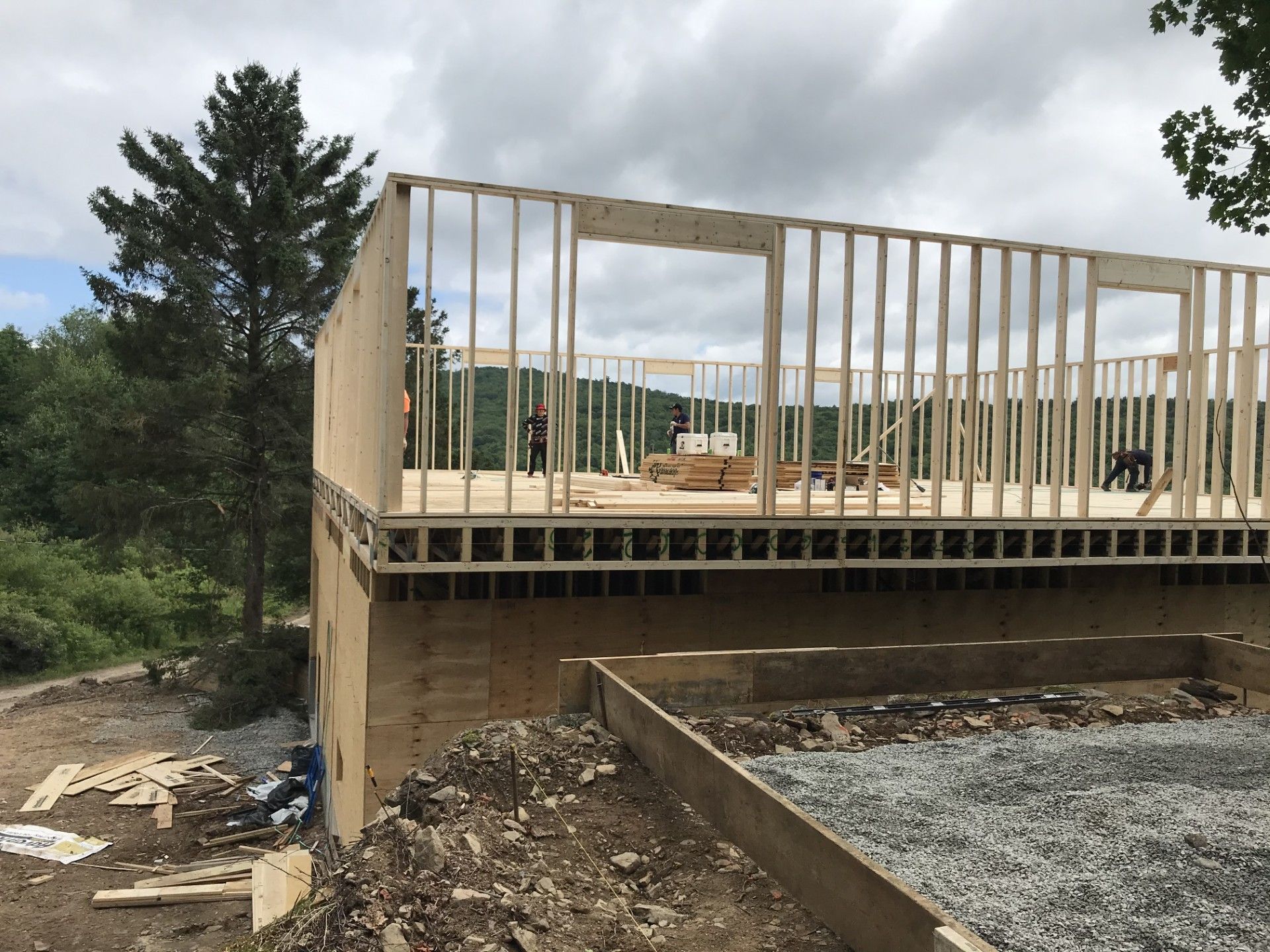 A wooden wall frame under construction on top of a foundation with trees and a cloudy sky in the background.