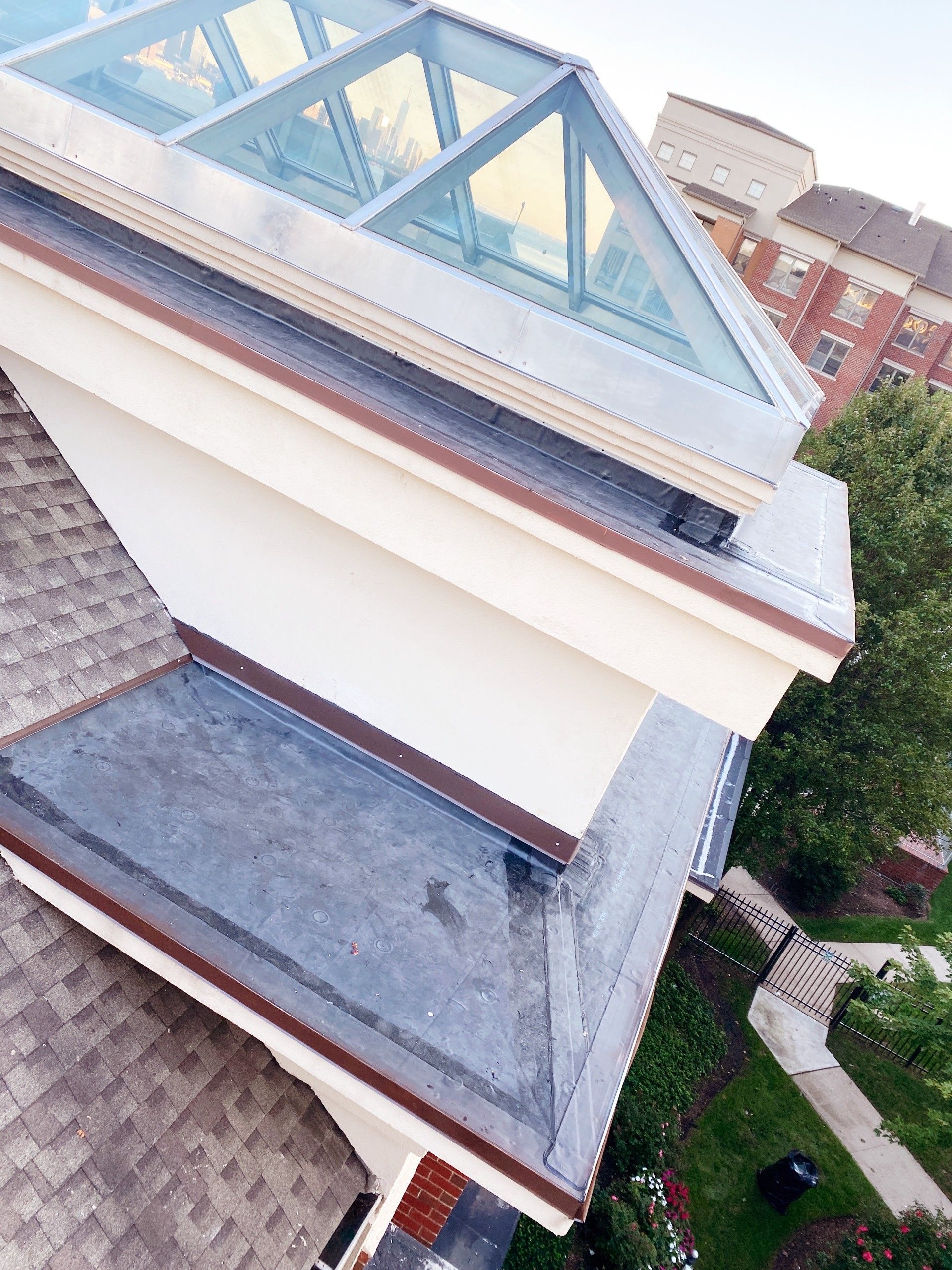 A high-angle view of a flat roof section featuring a large glass skylight, dark roofing material, and cream-colored trim.