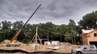 Construction site with a crane lifting materials, power lines overhead, a new house under construction, and a pickup truck.